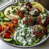 Vibrant falafel bowl features a generous drizzle of rich tahini sauce over fresh salad greens, cherry tomatoes, cucumber, and red onion. 
